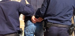 Policjanci prowadzą zatrzymanego mężczyznę z założonymi kajdankami na ręce z tyłu.
