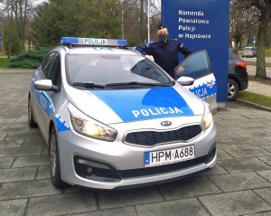 Policjant stoi przy radiowozie. W tle napis Komenda Powiatowa Policji w Hajnówce.