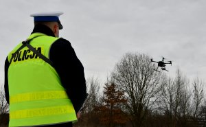 policjant obserwuje unoszącego się drona; w tle drzewa