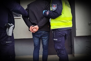 policjanci prowadzą osobę zatrzymaną, która ma założone kajdanki na ręce trzymane z tyłu